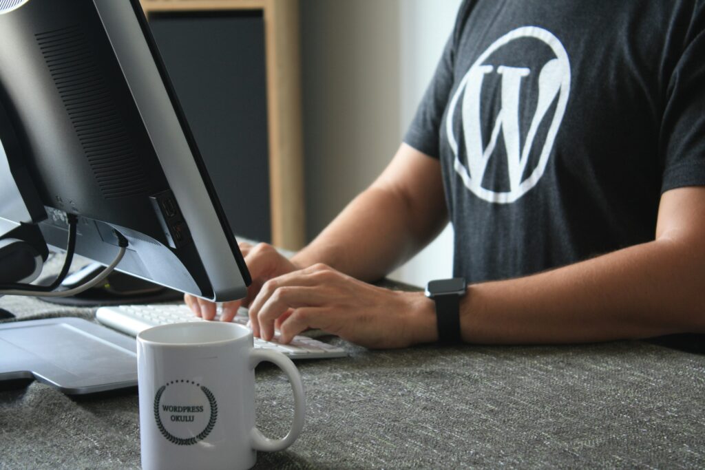 a person at a desktop computer wearing a black t-shirt with a white Wordpress logo on it.